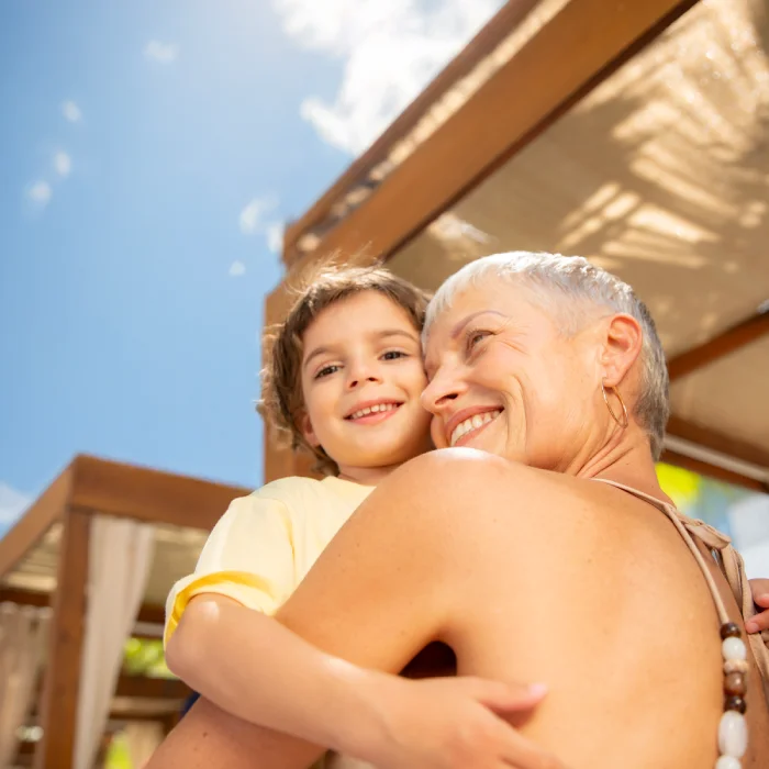 Short-haired woman affectionately hugging a young boy under a wooden pergola