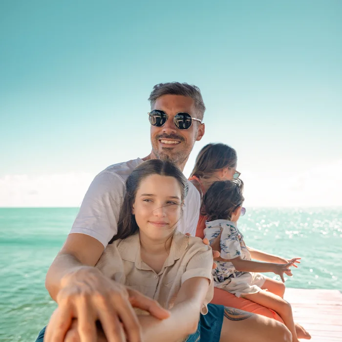 Man with sunglasses and his daughter sitting on a wooden pier facing the turquoise sea.